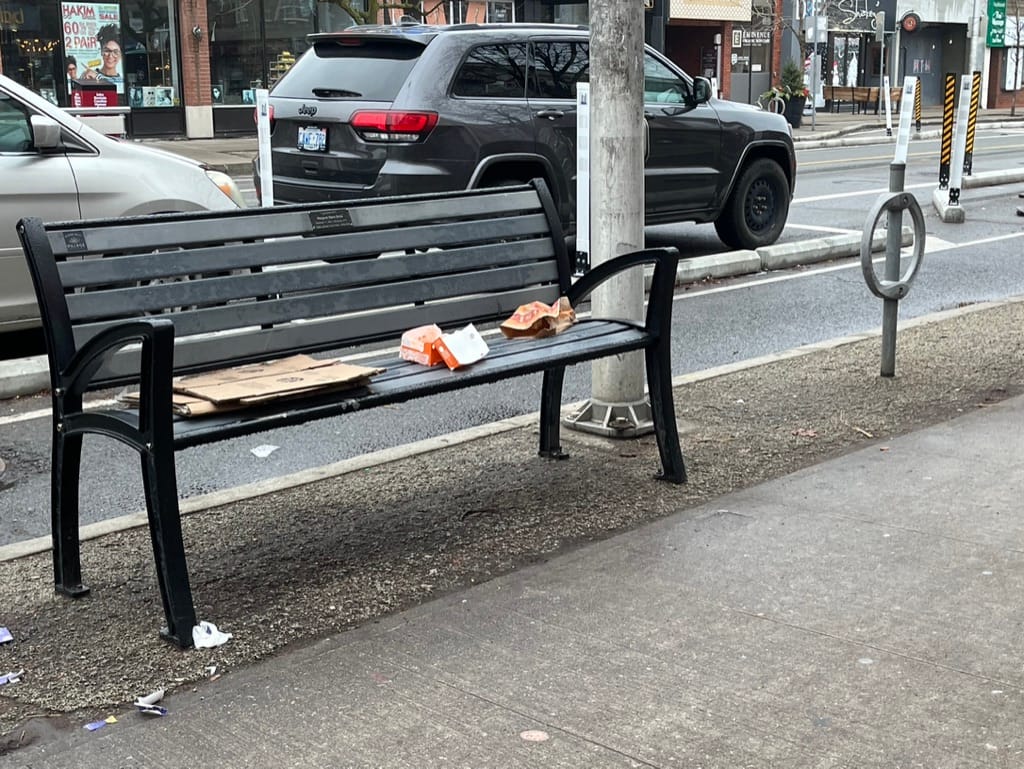 Image of a bench on Bloor Street West in Toronto. There are folded pieces of cardboard and empty fast food packaging on the bench.